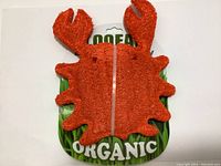 Front view of orange crab-shaped loofah dog toy on green packaging labeled Organic.