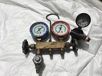 Dual manifold gauge set showing blue and red pressure dials, brass body, attached hoses and hanging hook, displayed on white cloth.