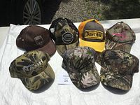 Seven ball caps arranged on a white cloth, featuring various camouflage patterns and branding embroidered on front panels. Caps are in like new condition.