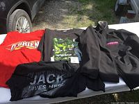 Four racing apparel items laid out on a table including a black Bicknell jacket and three t-shirts with various racing logos and graphics.