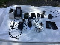 All cell phones, chargers, and cables laid out on white cloth outdoors showing variety and condition