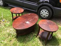 Photo of three wooden tables displayed outside on grass near a vehicle. The set includes one oval coffee table and two round end tables. The surfaces display visible wear expected from used furniture.