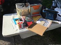 Wide view of crafting supplies laid out on a white folding table including tote bag, envelopes, papers, bead container and craft tools.