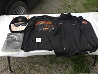 Full lot showing all four Harley Davidson items on white table outdoors: the plush backpack, graphic shirt, black jacket, and book.