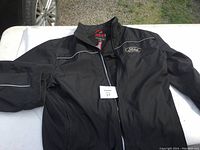 Front view of black Ford jacket with white piping detail and logo on chest, laid flat on surface.
