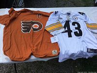 Two jerseys laid out on white surface; white Steelers jersey with number 43 on right, orange Flyers jersey with number 16 on left, front view.