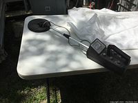 Full view of the metal detector showing the long metal shaft with circular detection coil and control box placed on white table outdoors.