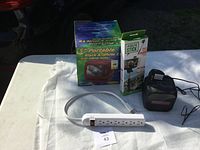 Full view of lot showing boxed vintage 5-inch black and white TV with AM/FM radio, selfie stick, digital clock radio, and power strip on white table outdoors.