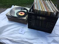 Photo showing a black plastic crate filled with vintage vinyl records and a pile of records including both 12-inch LPs and 7-inch singles on a white cloth.