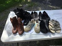 Seven pairs of used women's shoes and boots displayed on a white sheet outdoors on a table, showing all items in the lot