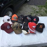 Photo of a collection of 10 sports ball caps laid out on a white table outdoors. Caps feature logos from Philadelphia Flyers, Boston Red Sox, and other sports teams in various colors and styles.