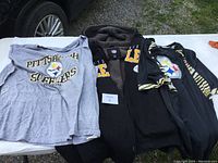 Three Pittsburgh Steelers clothing items laid out on a table including a black hoody coat, gray long sleeve shirt, and black jersey shirt, all size XXL.