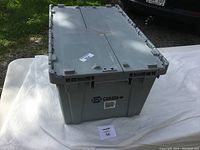 Closed grey plastic storage tote with hinged lid, labeled Dura Tuff Canada, placed on white surface outdoors.