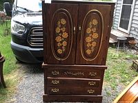 Photo of front view showing the wooden wardrobe with dark stain, two doors with floral painted oval panels, two drawers with brass-tone handles and carvings.
