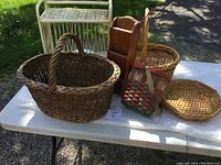 Five different wicker and wooden items on a white table including a large brown oval basket, two smaller round wicker baskets, a red and natural woven basket with a strap, and a brown wooden magazine rack.
