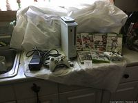 Photo of Xbox 360 console, two wireless controllers, power adapter, cables and 7 game cases arranged on a cloth-covered surface.