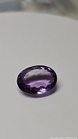 Single oval cut deep purple Brazilian amethyst gemstone placed on a light background showing faceted surface and color details.