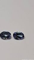 Two oval cut purple-blue Tanzanite gemstones with facet cuts visible, lying flat on white background showing color and clarity.
