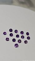 Fourteen natural faceted purple amethyst round stones laid out on a white background, showing diamond cut facets and deep purple color.