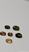 Six oval-cut natural tourmaline gemstones displayed on plain background showing variety of colors including strong orange, yellow, and greenish tones.