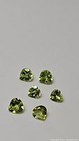 Six natural apple green peridot gemstones shown in a loose arrangement. Two heart-shaped and four teardrop-shaped stones visible with brilliant facets.