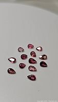 Photo of 13 pear-shaped natural pink tourmaline gemstones, showing varying shades of pink and faceted cuts.