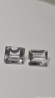 Two light-colored rose quartz gemstones shown side by side, emerald cut with clear transparency and light pink tint visible, measures approximately 10x8mm each.
