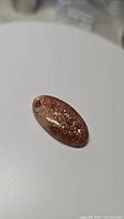 Oval natural sunstone cabochon photographed on white background, showing sparkly inclusions and polished surface.