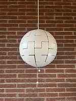 White IKEA PS 2014 spherical pendant lamp fully closed, hanging against a brick wall, showing the exterior closed panels.