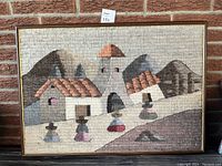 Framed tapestry showing entire village scene with church and figures, hung against brick wall