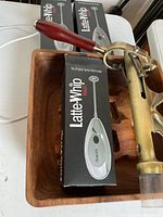 Top view of brass bottle corker with wooden handle and plastic sealing part, sitting inside wooden salad bowl alongside three boxed Latte Whip milk frothers.