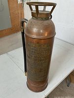 Full side view of the vintage copper fire extinguisher standing upright on a white surface, showing the hose attached to the side and the metal handle top.