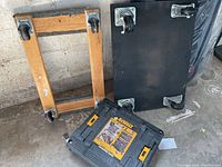 Three dollies arranged on concrete floor. Wooden dolly frame and black board dolly with four swivel casters each visible. DeWalt dolly cart with yellow latches visible.