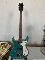 Full front view of Eastwood electric guitar showing metallic teal double-cutaway body, white pickguard, headstock and stand