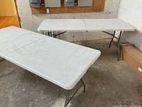 Two white folding tables positioned in a room, showing their tops and metal legs.