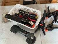 Full view of plastic basket containing various hand tools, 3 plastic spring clamps beside it, and a tool resembling a folding ruler or multi-tool.