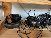 Photo showing a selection of black over-ear headphones, one marked 'GP-20 Electronic', with tangled cables on a wooden shelf