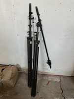 Two black adjustable equipment stands with counterweights and tripod bases, collapsed and leaning against a wall.