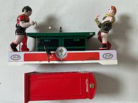 Top view of mechanical table tennis toy with two players on opposite ends of the green table; red metal telephone booth bank placed below.