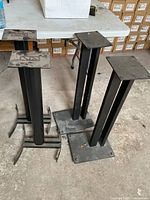 Four metal speaker stands in used condition with dust and surface dirt, showing different base styles.