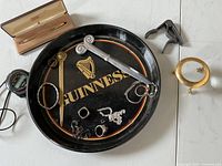 Photo of miscellaneous items on white surface including Cross pen in case, Guinness tray, rings, magnifying glass, candle holder, cutter and opener.