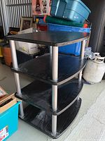 Multi-tiered table showing three black wood shelves supported by cylindrical metal legs. Side angle shows wear on surfaces and dusty condition.