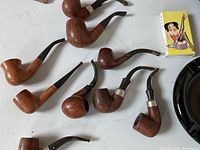 Overhead view of seven briar wood tobacco pipes on white surface with matchbox for scale