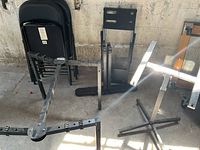 Photo showing four black metal folding chairs stacked and various collapsible metal stands with adjustable features and bases.
