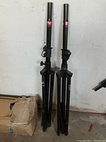 Two black, collapsible Yorkville SKS equipment stands seen standing against a white wall with folded tripod legs, approx. 48 inches collapsed height.