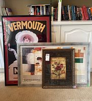 Three framed art prints displayed leaning against a white cabinet and bookshelf. Visible are Vermouth Martini poster, Maison Belle Jardiniere floral print, and a third framed piece behind them partially visible.