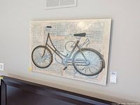 Frontal view of a bicycle print mounted on canvas with a newspaper-style text background hung on a light wall above a ledge.