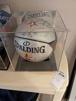 Front and side view of white Spalding All-Star basketball with visible signatures inside a clear acrylic case on a light-colored table.