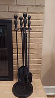 Set of four black metal fireplace tools including poker, tongs, broom, and shovel on a round metal stand with a brick fireplace in the background.