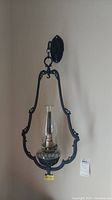 Full view photo of metal framed hanging oil lamp mounted on a wall, showing the black metal frame and glass chimney.